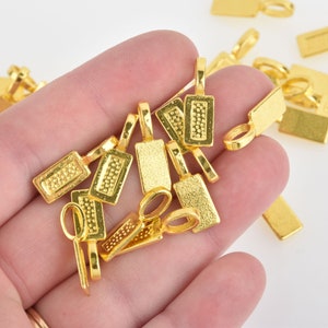 May include: A close-up of a hand holding a pile of gold-colored metal pendant charms. The charms are rectangular with a textured surface and a loop at the top.