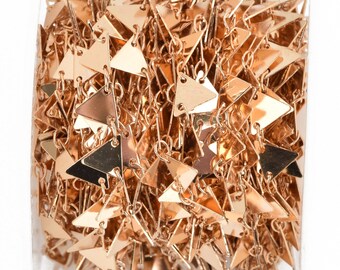 May include: Close-up of a roll of gold-colored chain. The chain is made of small, flat, triangle-shaped pieces linked together. The triangles are shiny and reflect light, creating a shimmering effect. The chain is tightly wound, showing the intricate design.