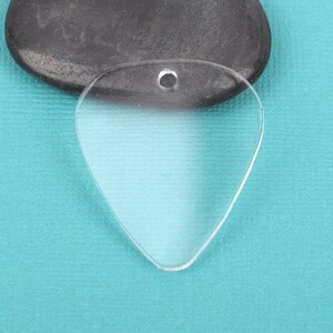 May include: A clear plastic guitar pick with a hole at the top for a string or chain. The pick is shaped like a traditional guitar pick and is lying on a blue surface.
