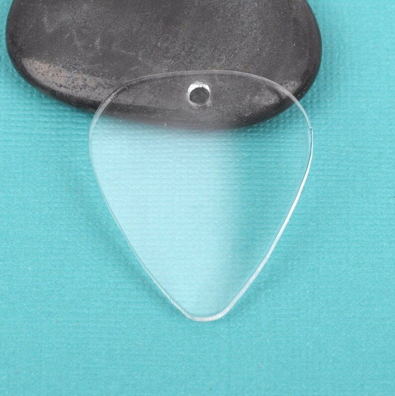 May include: A clear plastic guitar pick with a hole at the top for a string or chain. The pick is shaped like a traditional guitar pick and is lying on a blue surface.