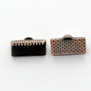 May include: Two small, rectangular metal clasps with a copper finish. One clasp has a textured, dotted surface and a scalloped edge. The other clasp has a dotted surface and a small loop on top. These clasps are likely used for jewelry making or crafts.