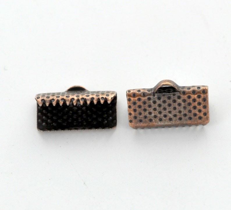 May include: Two small, rectangular metal clasps with a copper finish. One clasp has a textured, dotted surface and a scalloped edge. The other clasp has a dotted surface and a small loop on top. These clasps are likely used for jewelry making or crafts.