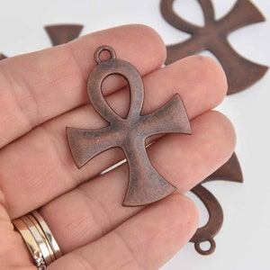 May include: Close-up of several bronze-colored Ankh charms. The charms are in the shape of a cross with a loop at the top. The charms have a rustic, antique finish. The charms are likely used for jewelry making or crafting.
