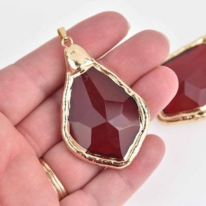 May include: A large, faceted, dark red glass pendant with a gold-toned metal frame. The pendant has a teardrop shape and is hanging from a gold-toned metal bail.