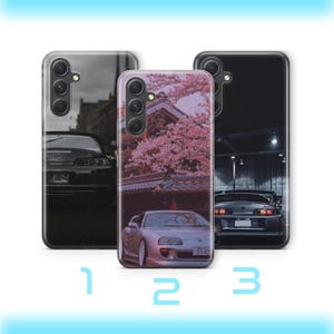 以下が含まれることがあります： 自動車のデザインが施された3つのスマートフォンケース。左のケースは暗い車の後部、中央は桜の下に駐車されたシルバーの車、右はガレージ内の車が描かれています。各ケースには3つのカメラレンズが配置されています。