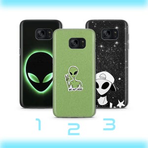 May include: Three phone cases with alien designs. One case is black with a glowing green alien head. Another is green with an alien giving a peace sign and the text "WE OUT HERE". The third case is black with a starry background and an alien wearing a hat.