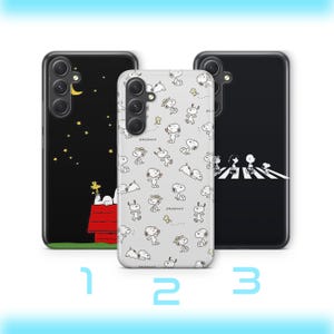 Op de afbeelding: Drie telefoonhoesjes met Snoopy-ontwerpen. Eén hoesje is zwart met een Snoopy- en Woodstock-scène onder een maan en sterren. Een andere is wit met Snoopy-personages. De derde is zwart met Snoopy die een straat oversteekt.