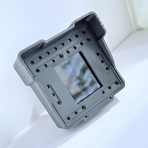 Puede incluir: Un dispositivo rectangular gris con una pantalla central reflectante. El dispositivo tiene múltiples agujeros pequeños y una superficie texturizada. El diseño incluye elementos redondeados y angulares, lo que sugiere un producto tecnológico o industrial.