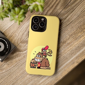 May include: Yellow phone case with a cartoon illustration of Donkey Kong and Diddy Kong. The design shows a brown gorilla holding a barrel with "DK" on it, and a monkey in a red hat with a banana. The case is on a wooden surface.