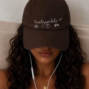 Puede incluir: Una gorra de béisbol de color marrón oscuro con el texto blanco Unstoppable en letra cursiva. Debajo del texto hay pequeños iconos de líneas blancas que representan una cámara, un corazón, un globo terráqueo, un todoterreno y un avión de papel.