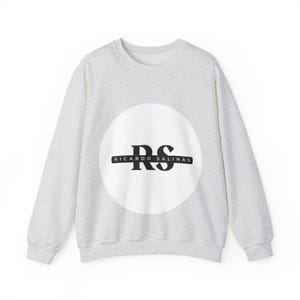 Puede incluir: Una sudadera de cuello redondo en color gris jaspeado claro con un gran círculo blanco en el pecho. Dentro del círculo hay una barra horizontal negra con las letras RS en fuente serif negrita y el texto RICARDO SALINAS en fuente sans-serif.