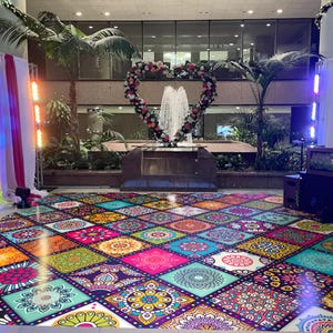 May include: A vibrant, colorful dance floor with a repeating pattern of square tiles, each featuring a unique mandala design in shades of pink, orange, blue, and green. A heart-shaped floral arrangement frames a fountain, creating a festive atmosphere.