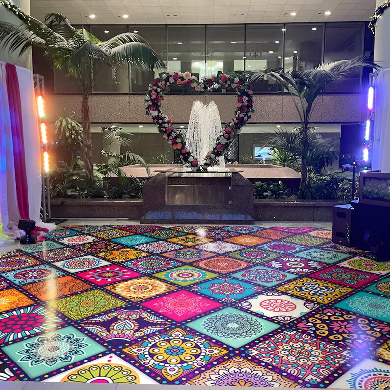 May include: A vibrant, colorful dance floor with a repeating pattern of square tiles, each featuring a unique mandala design in shades of pink, orange, blue, and green. A heart-shaped floral arrangement frames a fountain, creating a festive atmosphere.