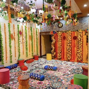 May include: A room decorated for a celebration, featuring floral arrangements and hanging garlands. The floor is covered with a patterned rug, and there are several small, round stools and cylindrical cushions. The walls are adorned with decorative elements, creating a festive atmosphere.