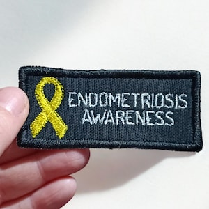 May include: A black embroidered patch featuring a yellow ribbon and the words "ENDOMETRIOSIS AWARENESS" in white. The patch is rectangular with a black border.