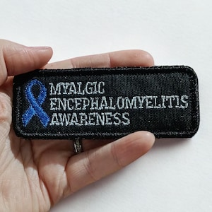 May include: A black rectangular patch with the text "MYALGIC ENCEPHALOMYELITIS AWARENESS" in white lettering. A blue ribbon graphic is on the left. The patch is held in a hand.