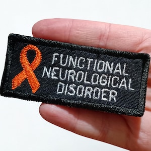 Peut inclure: Un écusson noir brodé avec les mots "FUNCTIONAL NEUROLOGICAL DISORDER" en blanc. Un ruban orange est sur le côté gauche de l'écusson. L'écusson est rectangulaire et a une bordure noire.