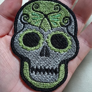 Op de afbeelding: Geborduurde schedelpatch met een grijze basis, zwarte oogkassen en een groen ontwerp op het voorhoofd. De patch is omrand met zwart en groen garen. Deze decoratieve patch kan op kleding of accessoires worden genaaid of gestreken.