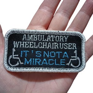 May include: Embroidered patch with the text "AMBULATORY WHEELCHAIR USER IT'S NOT A MIRACLE" in white and blue on a black background. The patch has a white border and features wheelchair symbols.