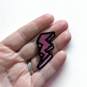 May include: A pink and black embroidered lightning bolt patch. The patch features a pink lightning bolt design with a black border. The patch is held in a person's hand against a white background. This patch can be ironed on to clothing.