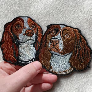 Op de afbeelding: Twee geborduurde patches met hondenportretten. De honden hebben bruine en witte vacht, met gedetailleerde steken. De patches hebben de vorm van de kop en schouders van de hond, met een zwarte rand.