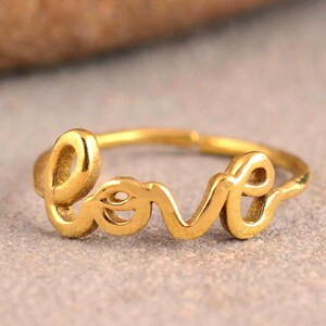 May include: A gold-toned metal ring featuring the word love written in a continuous cursive script. The band is thin and circular, with the letters forming the focal point of the design. The metal has a smooth, polished finish throughout the piece.