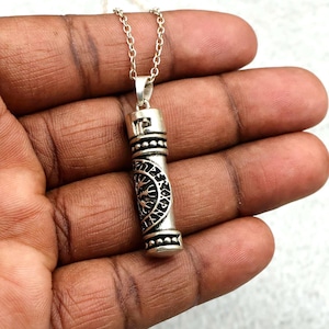 May include: A silver-toned cylindrical pendant necklace featuring an engraved Vegvisir symbol surrounded by runic characters. The pendant is suspended from a delicate silver-toned chain, showcasing a metallic finish with dark oxidised detailing.