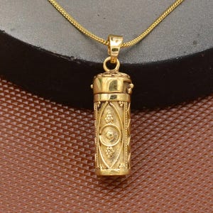 Puede incluir: Un collar con colgante dorado con un relicario cilíndrico con diseños en relieve intrincados. El relicario cuelga de una delicada cadena dorada. El colgante presenta un patrón detallado con elementos florales y geométricos.