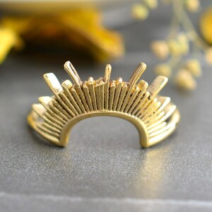 Puede incluir: Un anillo de tono dorado con una banda semicircular que presenta una serie de puntas verticales radiantes de alturas variables a lo largo del borde superior, creando un patrón de rayos de sol. La superficie metálica es lisa y pulida.