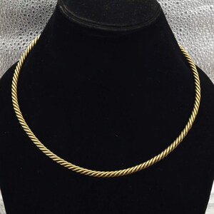 May include: A gold-toned choker necklace with a twisted rope design. The necklace is displayed on a black jewelry bust against a silver background. The choker has a simple, elegant style.
