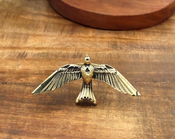 May include: A metallic gold-toned bird-shaped ring featuring outstretched wings and a small triangular cutout on the chest. The bird is depicted in a flying position with detailed feather engravings along the wings and tail, creating a textured surface.