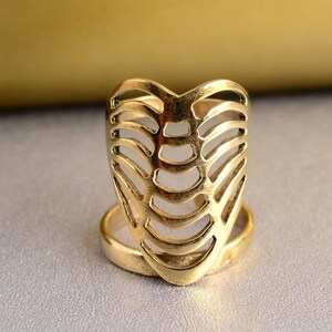 May include: A gold ring with a distinctive design. The ring has a cage-like structure, resembling a rib cage, with curved bars forming an openwork pattern. The ring sits on a plain band, and the overall style is striking and unusual.
