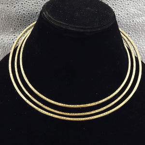 May include: A gold-colored necklace with three circular bands. The bands have a textured surface and are displayed on a black mannequin. The background is a light, textured fabric.