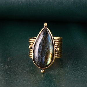 Pode incluir: Um anel em tom de latão com uma pedra de labradorita em forma de gota. A pedra exibe tons iridescentes de azul, verde e amarelo. A banda do anel tem um design canelado com pequenos detalhes redondos. O anel está sobre um fundo verde escuro.