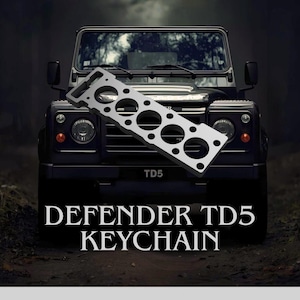 Peut inclure: Un porte-clés Defender TD5 argenté est présenté devant un Land Rover Defender noir. Le porte-clés a une forme unique avec plusieurs découpes circulaires. Le texte "DEFENDER TD5 KEYCHAIN" est en bas.