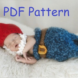 May include: A baby wearing a red knitted hat with a white beard and a blue knitted gnome costume with a brown belt and a yellow buckle. The baby is lying on a white fluffy surface. The text "PDF Pattern" is written in blue letters at the top of the image.