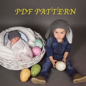 May include: Two children wearing gray bunny hats with white ears. The younger child is sleeping in a white wicker basket with pastel colored Easter eggs. The older child is sitting on the floor holding a white speckled Easter egg.  PDF PATTERN.