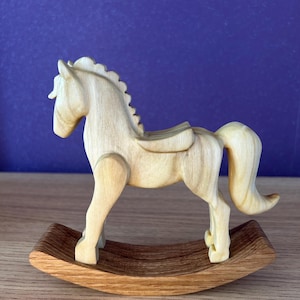 May include: A light-coloured wooden rocking horse figurine featuring a smooth, carved body with a defined mane, tail, and saddle. The horse is mounted on a curved, dark-toned wooden rocker base, showcasing natural wood grain patterns throughout the piece.