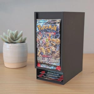 May include: A black, rectangular card holder with two compartments. The top compartment holds a Pokemon trading card pack with the text "Pokemon Trading Card Game Stellar Crown". The bottom compartment holds additional card packs. A succulent plant in a white pot is to the left.