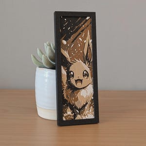 May include: A framed art piece featuring a cartoon Eevee character in shades of brown and beige. The artwork is set against a snowy background and framed in a dark rectangular border. A small succulent plant in a white pot is next to the artwork.
