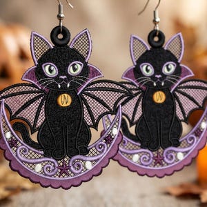 May include: A pair of embroidered earrings featuring black cats with bat wings, vampire fangs, and purple collars. Each cat sits on a crescent moon decorated with purple swirls, small stars, and white accents, suspended from silver-toned metal hooks.