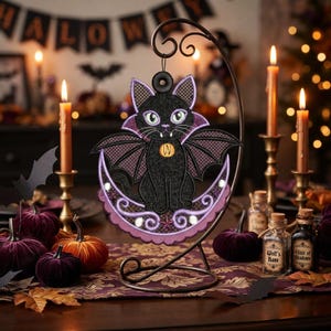 May include: A black cat vampire ornament with purple wings and a crescent moon base, hanging from a dark metal stand. The cat has green eyes, white fangs, and a small orange pumpkin charm around its neck, featuring detailed embroidery stitching.