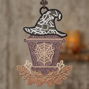 Op de afbeelding: Een hangend geborduurd ornament met een paarse lantaarn met spinnenwebpatroon, bekroond met een zwart-witte heksenhoed. De basis is versierd met lichtbruine geborduurde eikenbladeren en vier eikels in een symmetrische opstelling.