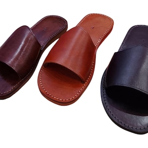 May include: Three leather slide sandals in shades of brown and black. The sandals have a simple design with a single strap across the top and are stitched around the edges. The leather appears smooth and polished.