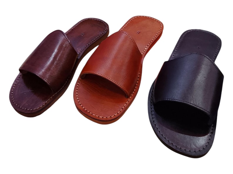 May include: Three leather slide sandals in shades of brown and black. The sandals have a simple design with a single strap across the top and are stitched around the edges. The leather appears smooth and polished.