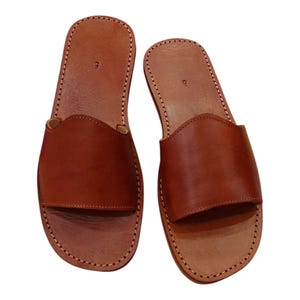 May include: A pair of brown leather slide sandals. The sandals have a simple design with a strap across the top and are stitched around the edges. The number "9" is visible on the footbed.