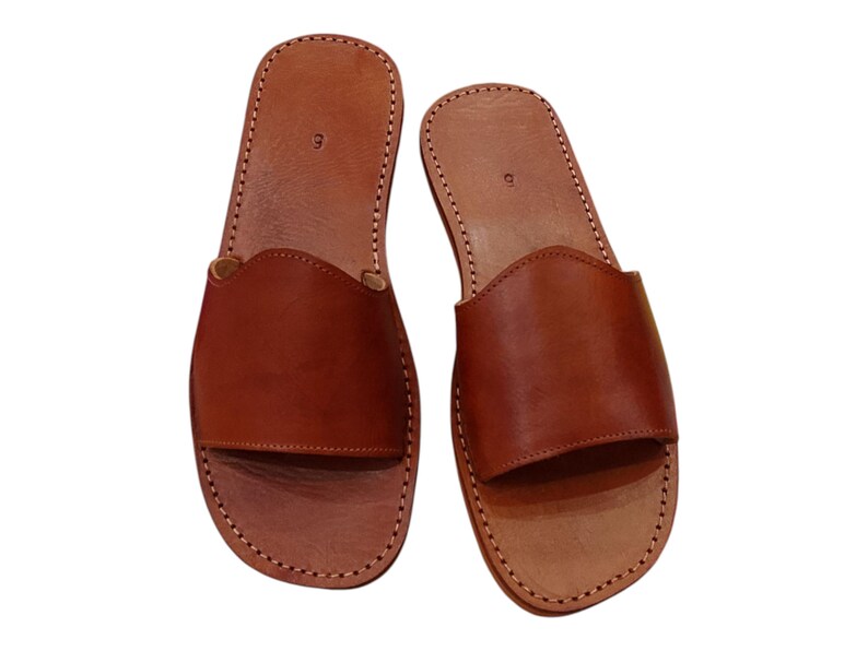 May include: A pair of brown leather slide sandals. The sandals have a simple design with a strap across the top and are stitched around the edges. The number "9" is visible on the footbed.