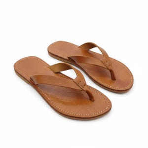 May include: A pair of brown leather flip-flops. The sandals feature a simple thong strap design and stitched soles. The leather has a natural, textured appearance. The flip-flops are displayed against a white background.
