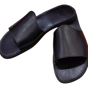 May include: A pair of black leather slide sandals. The sandals have a simple design with a wide strap across the top of the foot and a stitched sole. The sandals are made of a smooth, dark material.