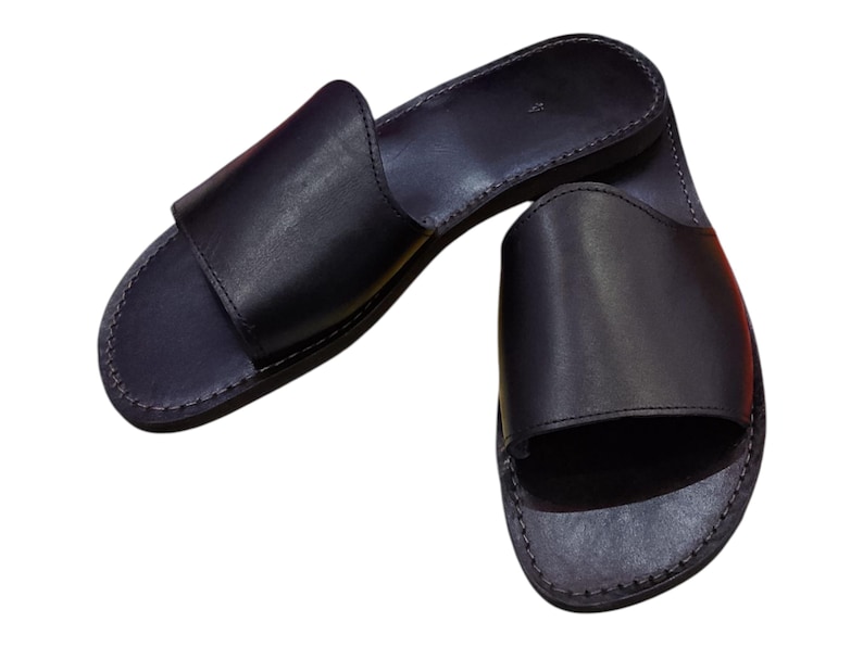 May include: A pair of black leather slide sandals. The sandals have a simple design with a wide strap across the top of the foot and a stitched sole. The sandals are made of a smooth, dark material.
